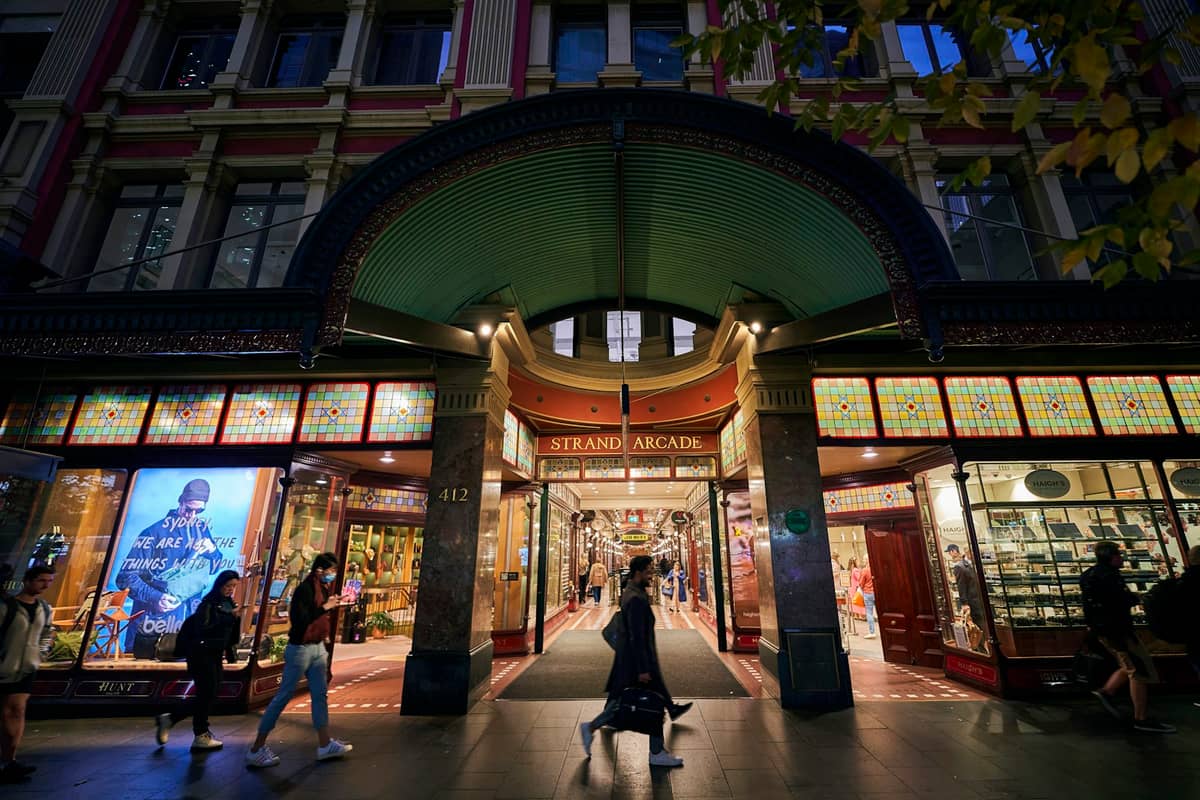 Abendlicher Blick auf den Haupteingang der Strand Arcade mit Buntglas-Beschilderung und Passanten vor den Schaufenstern der Boutiquen.