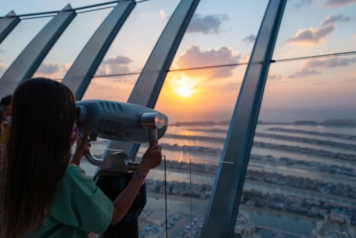 Im Inneren der Aussichtsplattform The View at The Palm schaut eine Person durch ein Fernglas auf den Sonnenuntergang über dem Palm Jumeirah und dem Arabischen Golf.