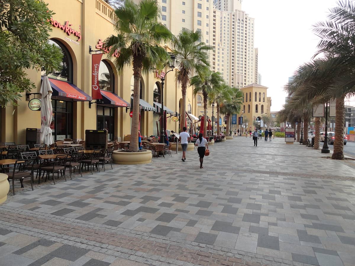 The Walk at JBR in Dubai mit einer breiten, palmengesäumten Fußgängerpromenade, Außensitzplätzen, flanierenden Menschen und hohen Wohntürmen im Hintergrund.