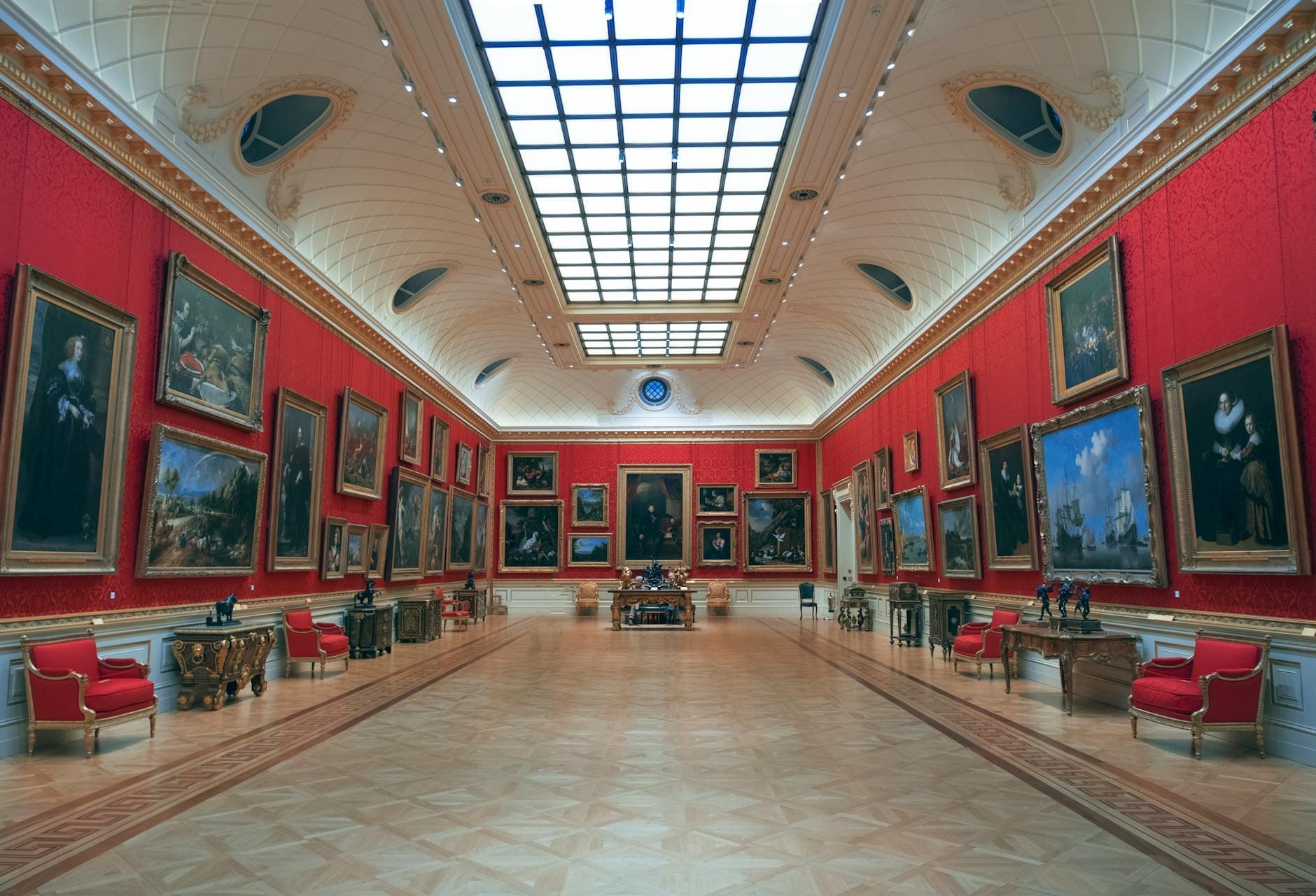 Una grande e sontuosa sala della Wallace Collection a Londra, con pareti rosse coperte di dipinti di maestri antichi, mobili ornati e un soffitto di vetro luminoso.
