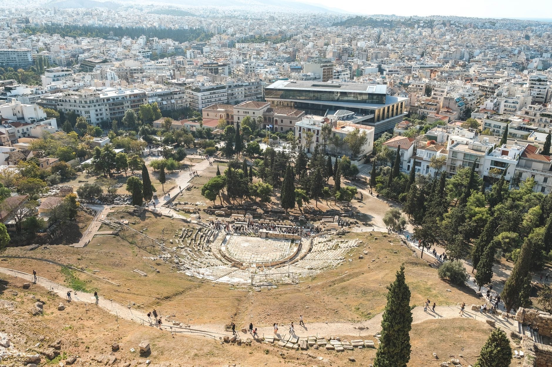 Vista aerea dell'antico Teatro di Dioniso Eleutereo sul versante dell'Acropoli, circondato dalla moderna Atene e da alberi verdi sotto un cielo sereno.