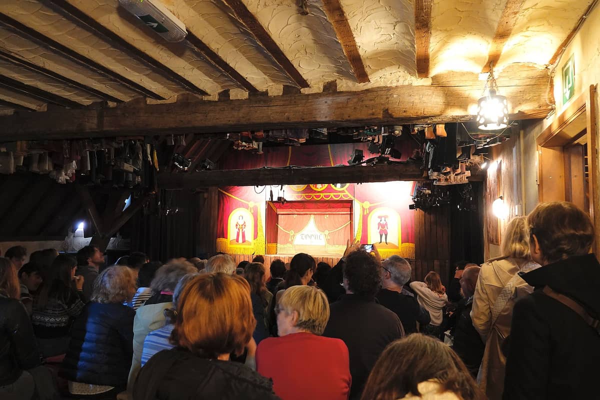Plateia sentada num sótão à luz de velas, com vigas à mostra, assistindo ao palco de marionetes do Théâtre Royal de Toone em Bruxelas.