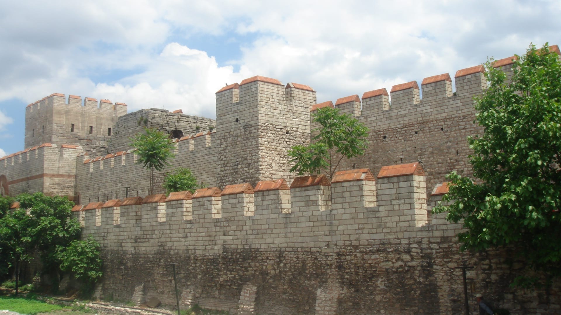 Bastioni e torri in pietra delle Mura Teodosiane a Istanbul sotto un cielo parzialmente nuvoloso, circondati da vegetazione.