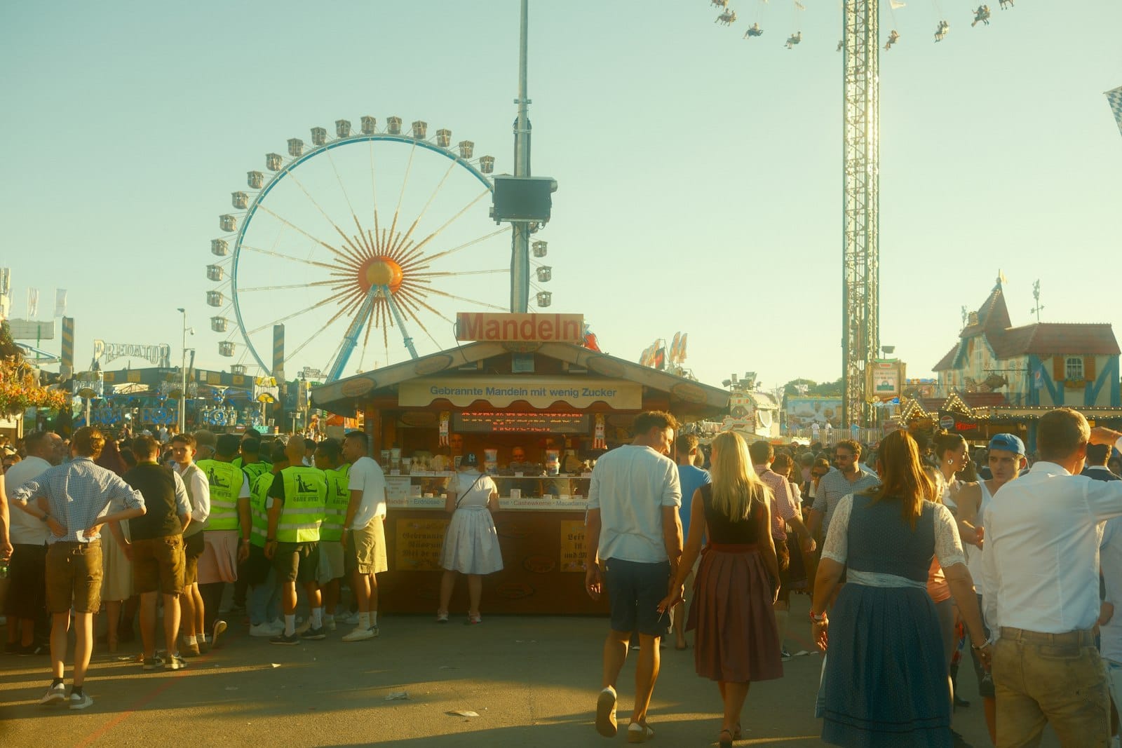ฝูงชนในชุดพื้นเมืองบาวาเรียเดินในลาน Oktoberfest ที่ Theresienwiese มิวนิก มีชิงช้าสวรรค์และร้านอาหารยามบ่ายแก่ๆ ท่ามกลางแสงแดดอุ่น