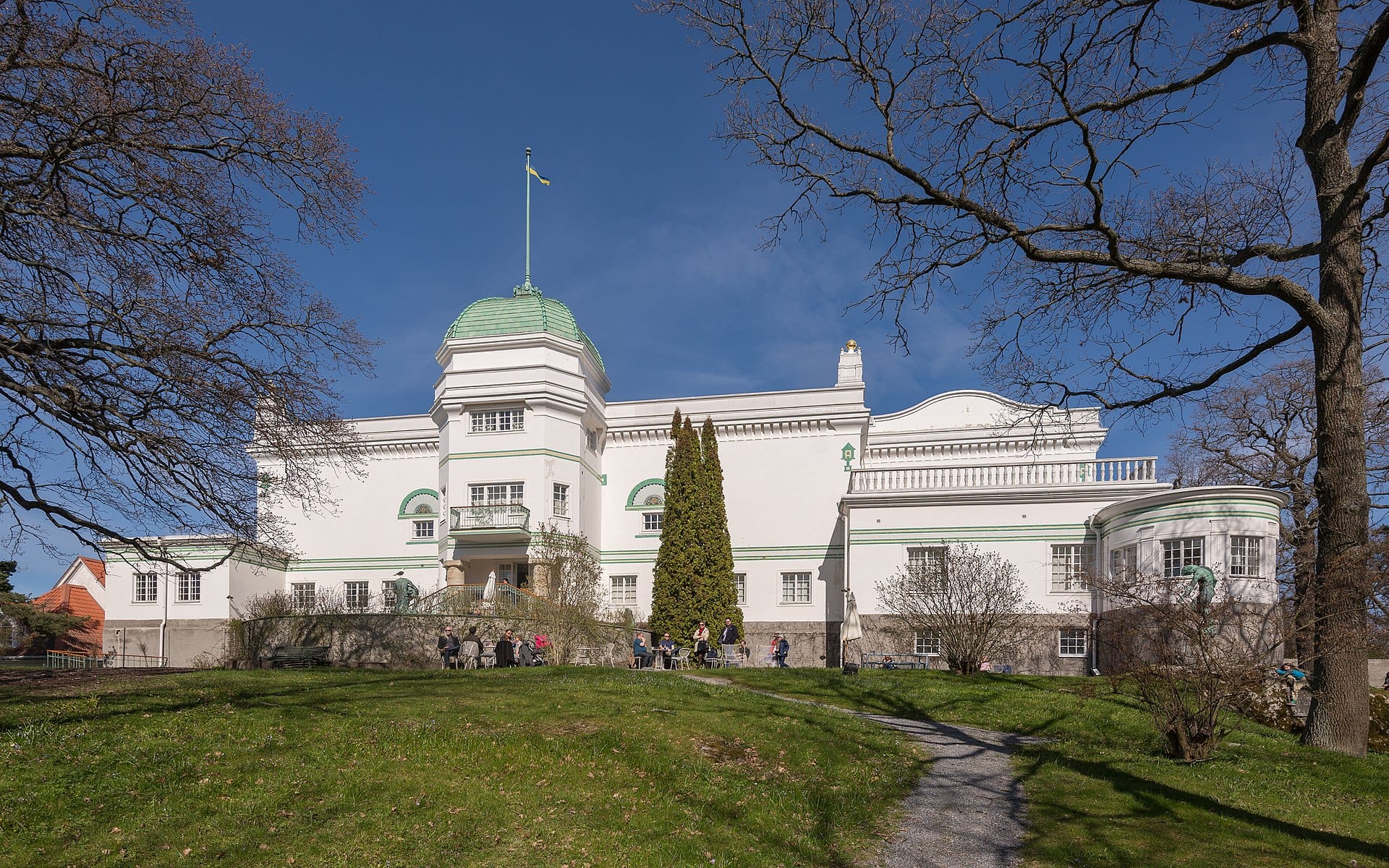 ภาพภายนอก Thielska Galleriet วิลล่าสีขาวสง่าหลังคาโดมเขียว มีธงชาติสวีเดน รายล้อมด้วยต้นไม้และผู้คนนั่งบนสนามหญ้า