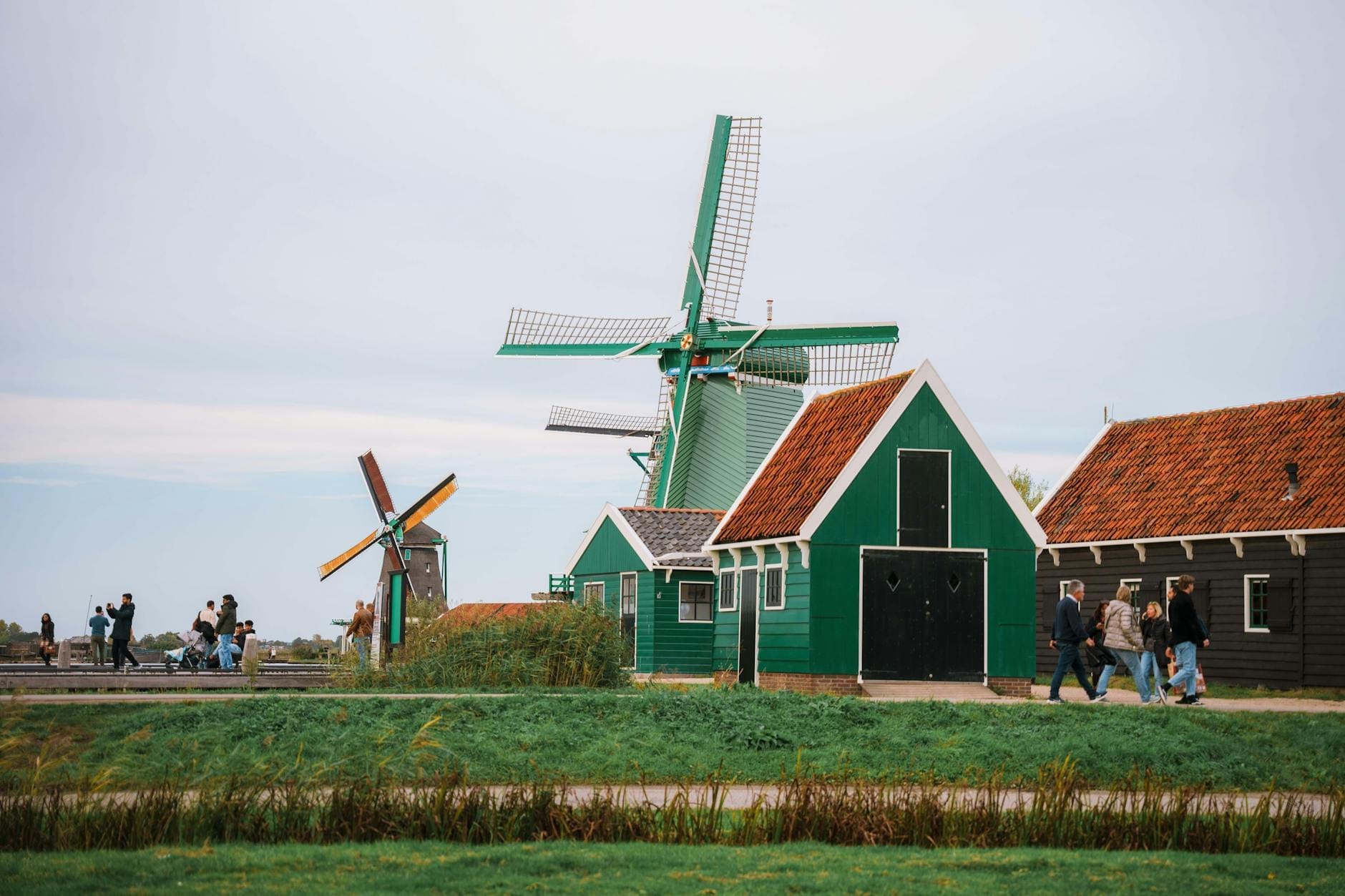 Turistas caminando junto a casas de madera verdes tradicionales y molinos de viento en funcionamiento cerca del agua, típicos de la zona de Zaanse Schans cerca de Ámsterdam.