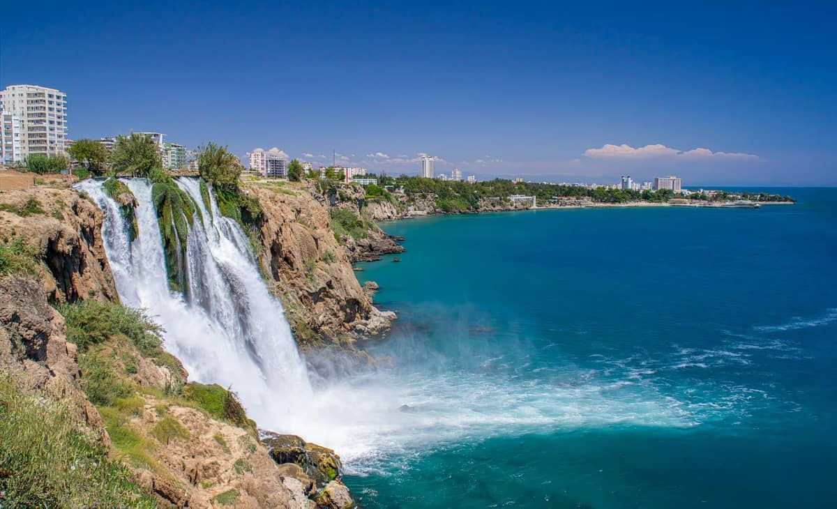 Düden Waterfalls cascading off cliffs into the Mediterranean Sea with Antalya city buildings and coastline in the background.