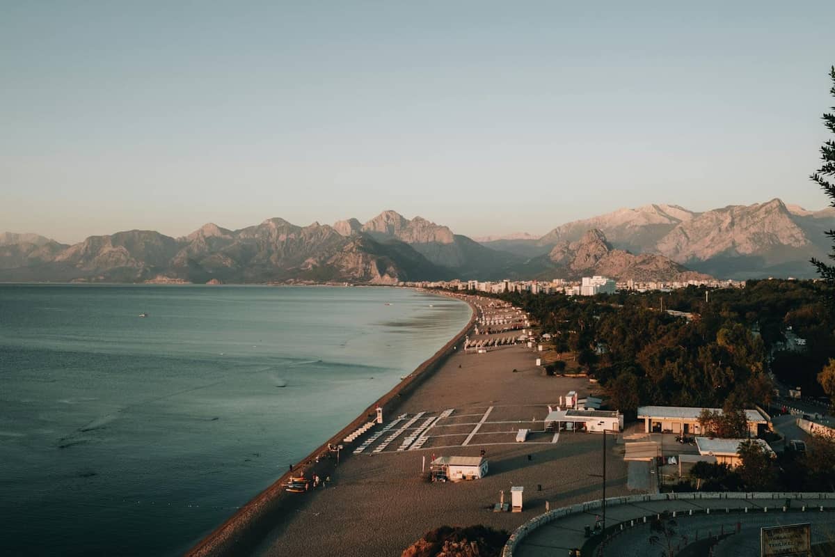 Wide view of Antalya beach with a long shoreline, sunbeds, turquoise water, city buildings, and mountains in the background.