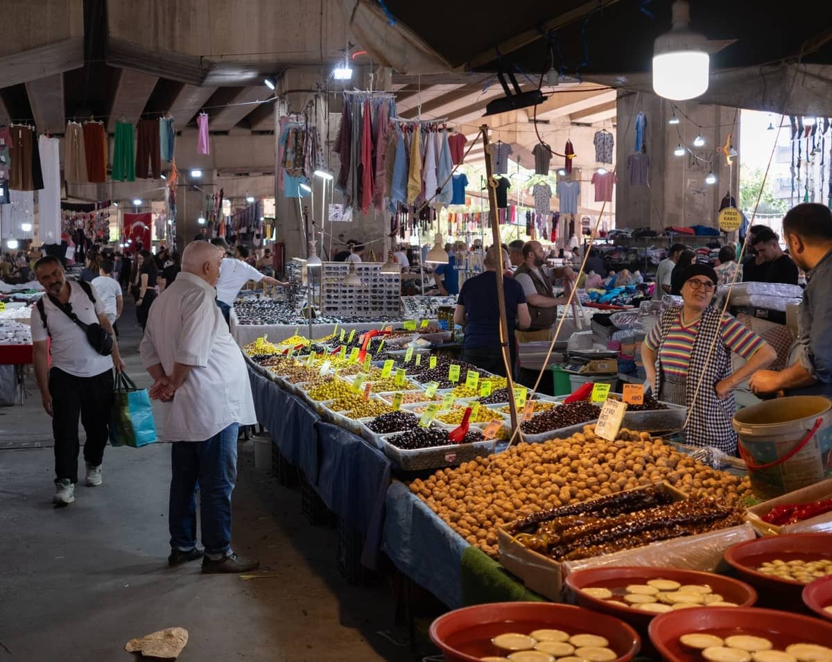Escena de mercado cubierto en Atenas con vendedores que ofrecen aceitunas, frutos secos y productos frescos, y personas comprando en varios puestos bien iluminados.