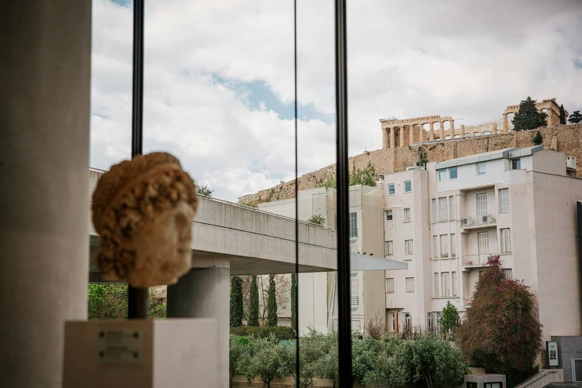Interior de un museo con una cabeza de estatua en primer plano y una vista por la ventana de la Acrópolis en Atenas