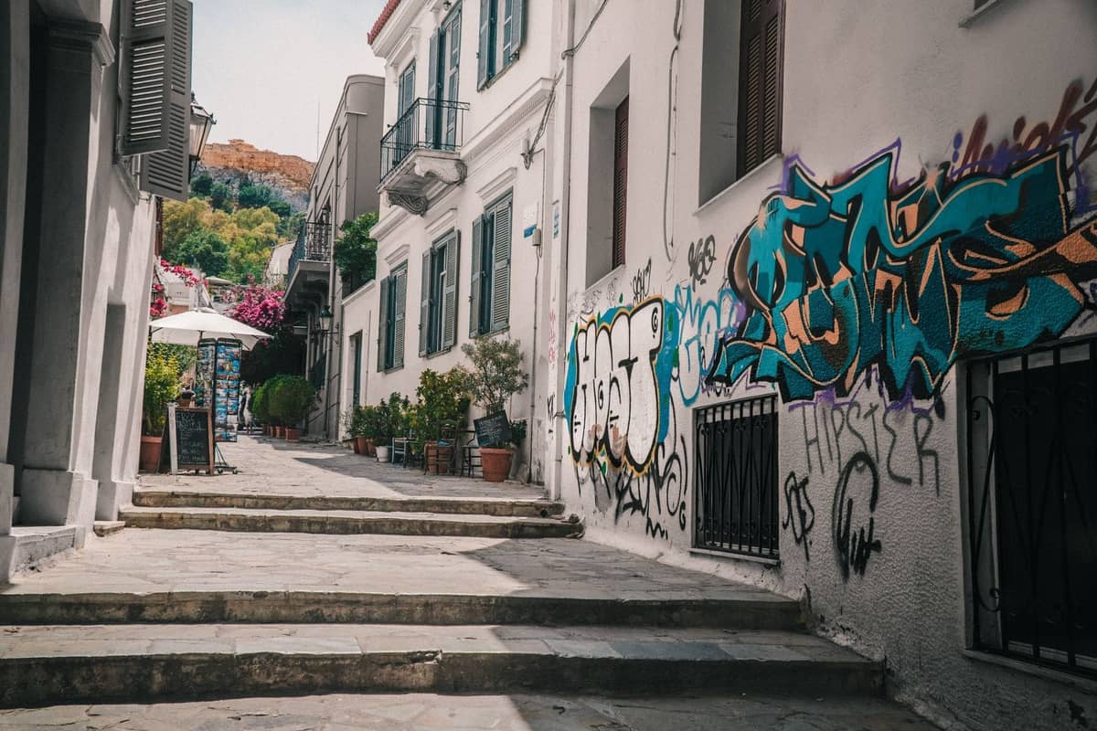 Calle peatonal estrecha en Atenas con casas encaladas, grafitis en las paredes, macetas y buganvillas, que sube por un animado barrio.