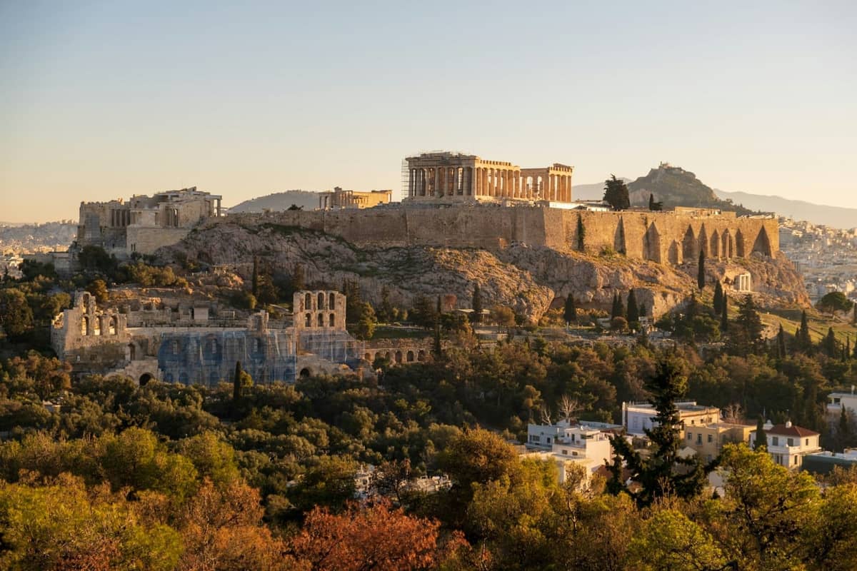 Vista panorámica de la colina de la Acrópolis en Atenas durante la hora dorada, con el Partenón, los Propileos, el Odeón de Herodes Ático y la vegetación circundante.