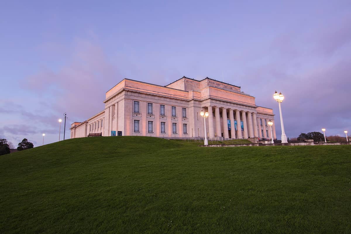 Un grandioso edificio de museo neoclásico con columnas, visto desde una colina cubierta de hierba al atardecer, con lámparas encendidas bajo un cielo pastel.