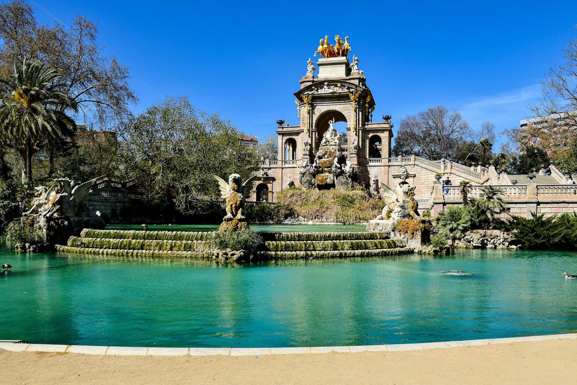 Wide view of the Cascada Monumental fountain and lake in Ciutadella Park, Barcelona, under a bright blue sky.
