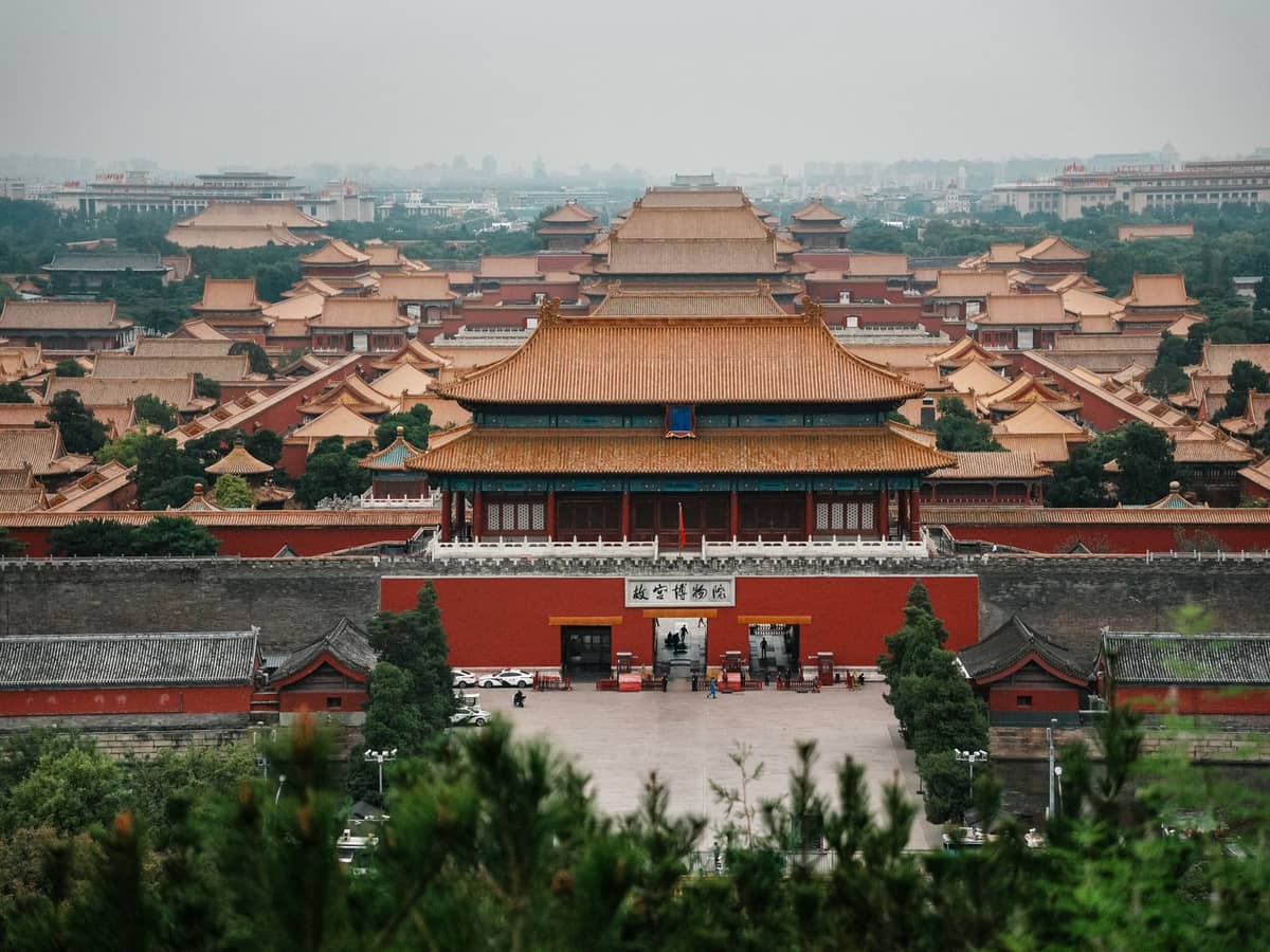 Weitläufiger Panoramablick auf die Verbotene Stadt in Peking mit goldenen Dächern und roten Mauern, umgeben von Grünflächen und der Stadtsilhouette im Hintergrund.