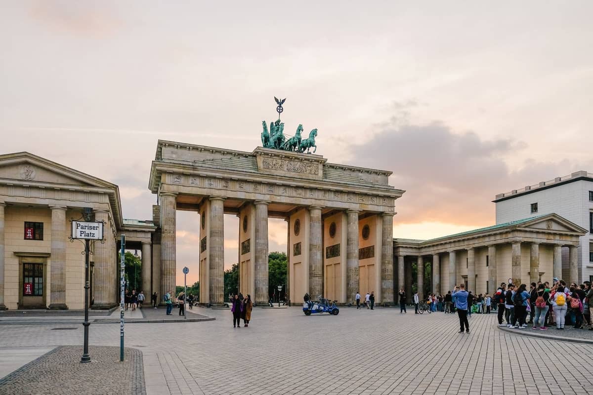 Abendlicher Blick auf das Brandenburger Tor in Berlin mit Touristen auf dem Platz und einem farbenfrohen Sonnenuntergang im Hintergrund.