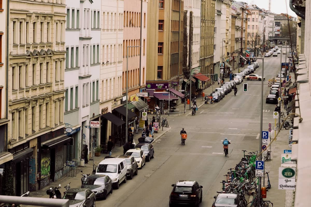 Eine lebhafte Berliner Straßenszene von oben mit bunten Gebäuden, Geschäften, Fahrrädern, geparkten Autos und Fußgängern und Radfahrern.