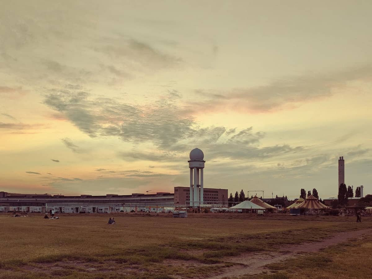 Tempelhofer Feld bei Sonnenuntergang, mit Menschen, die sich auf dem Rasen entspannen, und dem historischen Terminal im Hintergrund unter einem dramatischen Himmel.