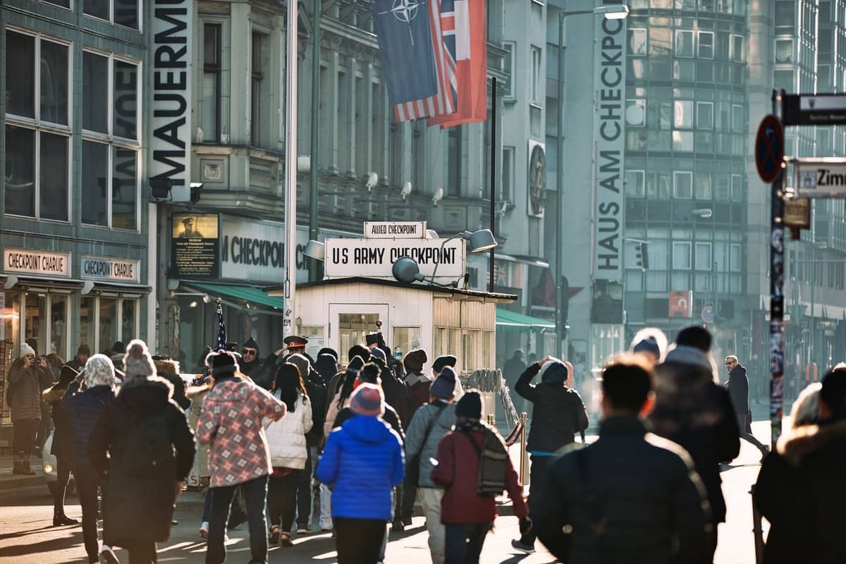 Touristen versammeln sich am historischen Checkpoint Charlie in Berlin, mit umliegenden Gebäuden und Beschilderung im Hintergrund.