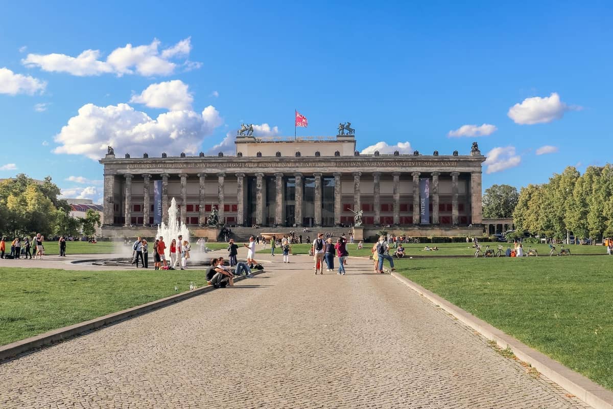 Weitwinkelansicht des Altes Museums auf der Berliner Museumsinsel mit Menschen, die sich auf grünen Rasenflächen entspannen, und einem Brunnen davor an einem sonnigen Tag