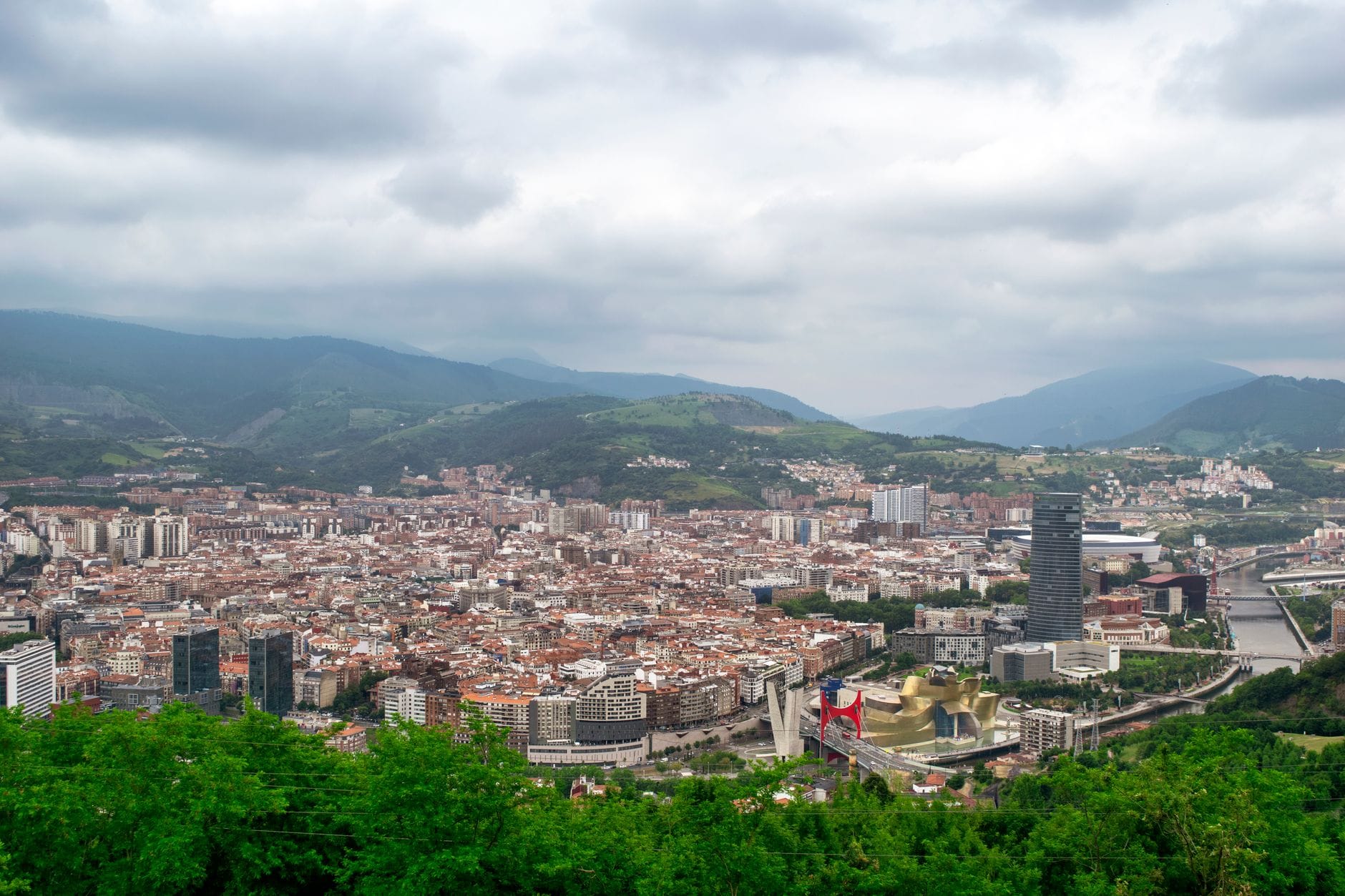 Panoramiczny widok dzienny na miasto Bilbao z zielonymi wzgórzami w tle, ukazujący miejską nieckę z wysokiego punktu widokowego.