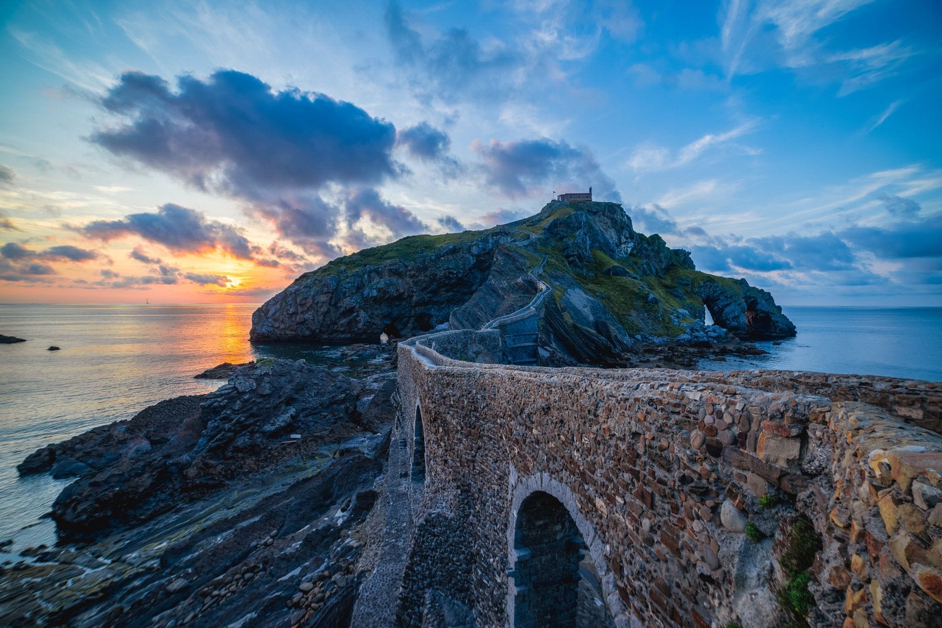 Kamienny most i schody prowadzące do pustelni na szczycie wysepki San Juan de Gaztelugatxe o zachodzie słońca na wybrzeżu Zatoki Biskajskiej.
