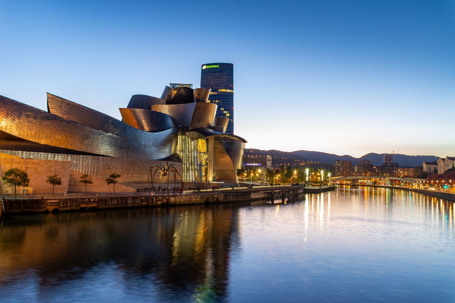 Szeroki widok na Guggenheim Museum Bilbao nad rzeką Nervión o zmierzchu, ze skyline miasta i nadrzeczną promenadą.