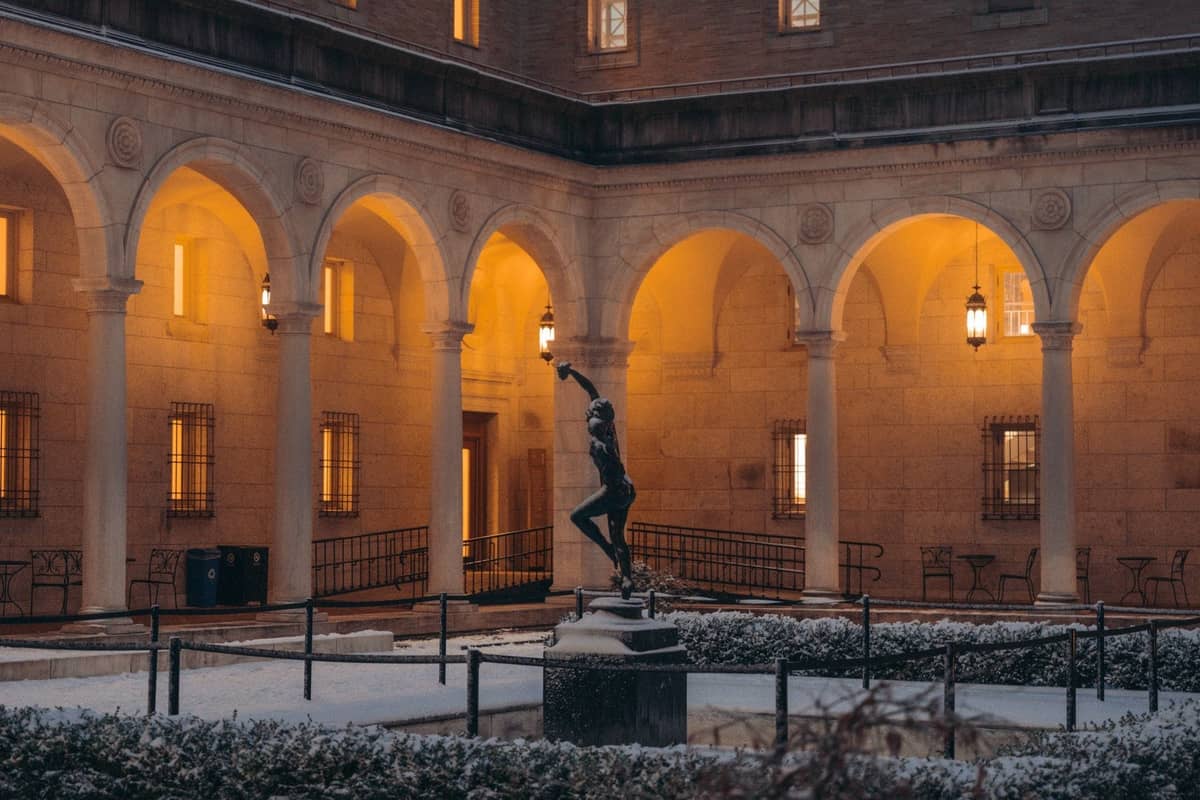 Cortile di un museo di Boston al crepuscolo, con archi, lanterne luminose e una statua al centro spolverata di neve.