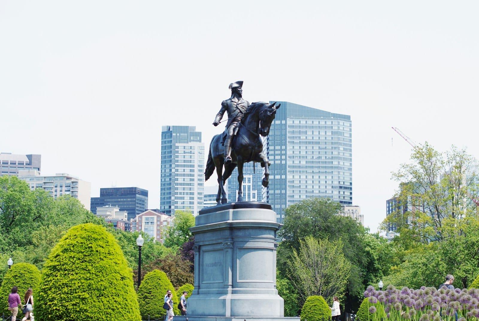 A estátua de George Washington no Boston Public Garden rodeada de arbustos bem cuidados e edifícios modernos da cidade num dia luminoso.