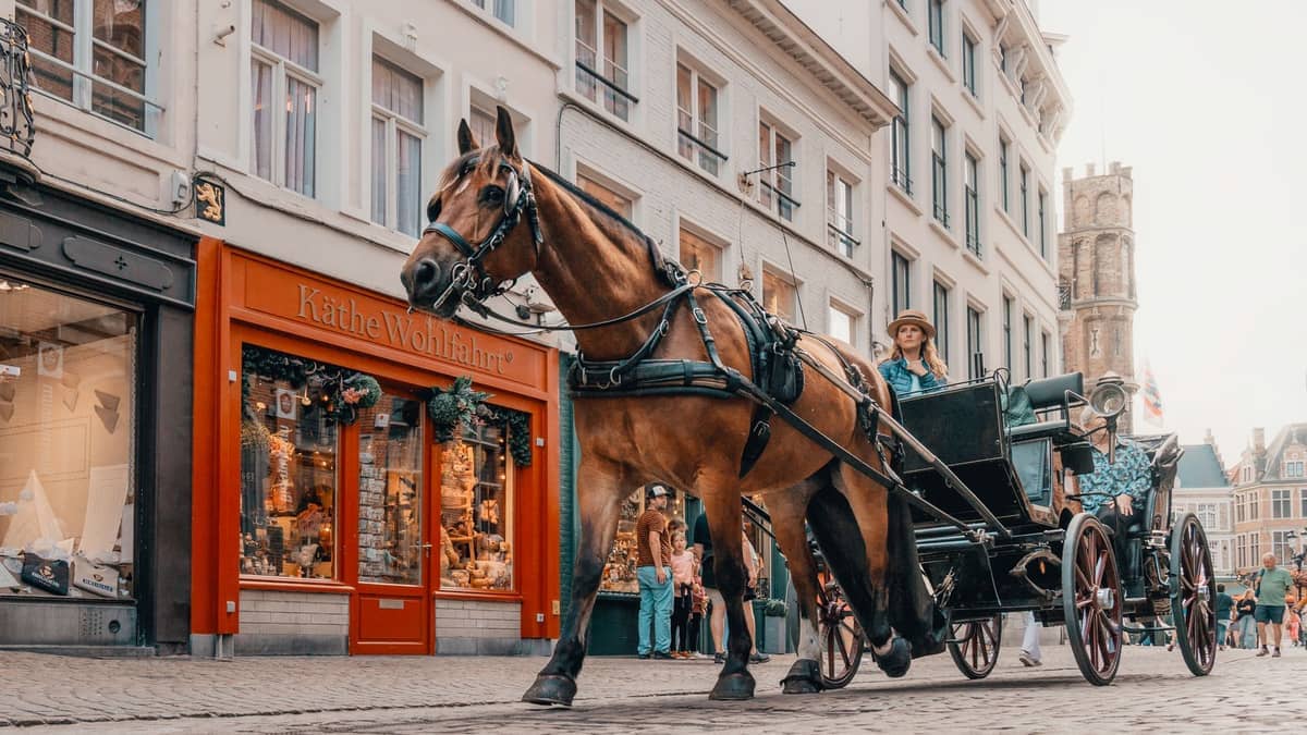 Horse-drawn carriage passing Käthe Wohlfahrt shop on a cobblestone street in Bruges, with pedestrians and medieval buildings in the background.