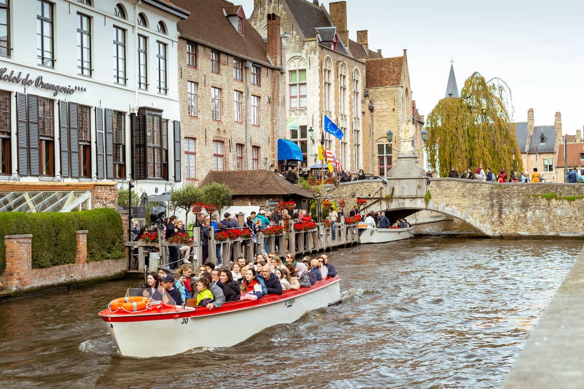 Tour boat full of passengers gliding on a Bruges canal, with historic waterfront buildings and a lively quay in the background.