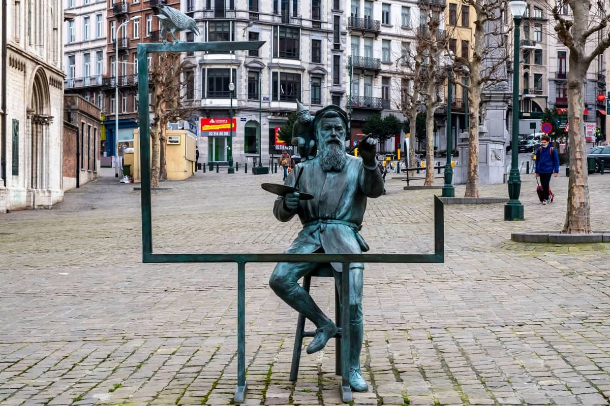 Estátua de bronze de um pintor com uma moldura na Place du Grand Sablon, Bruxelas, rodeada de edifícios históricos e árvores sem folhas no inverno.