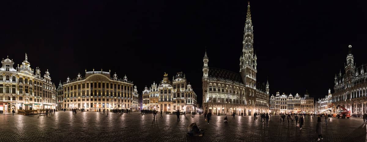 Vista panorâmica noturna da Grand-Place de Bruxelas, com as casas das guildas iluminadas e a Câmara Municipal gótica, animada com pessoas na praça.