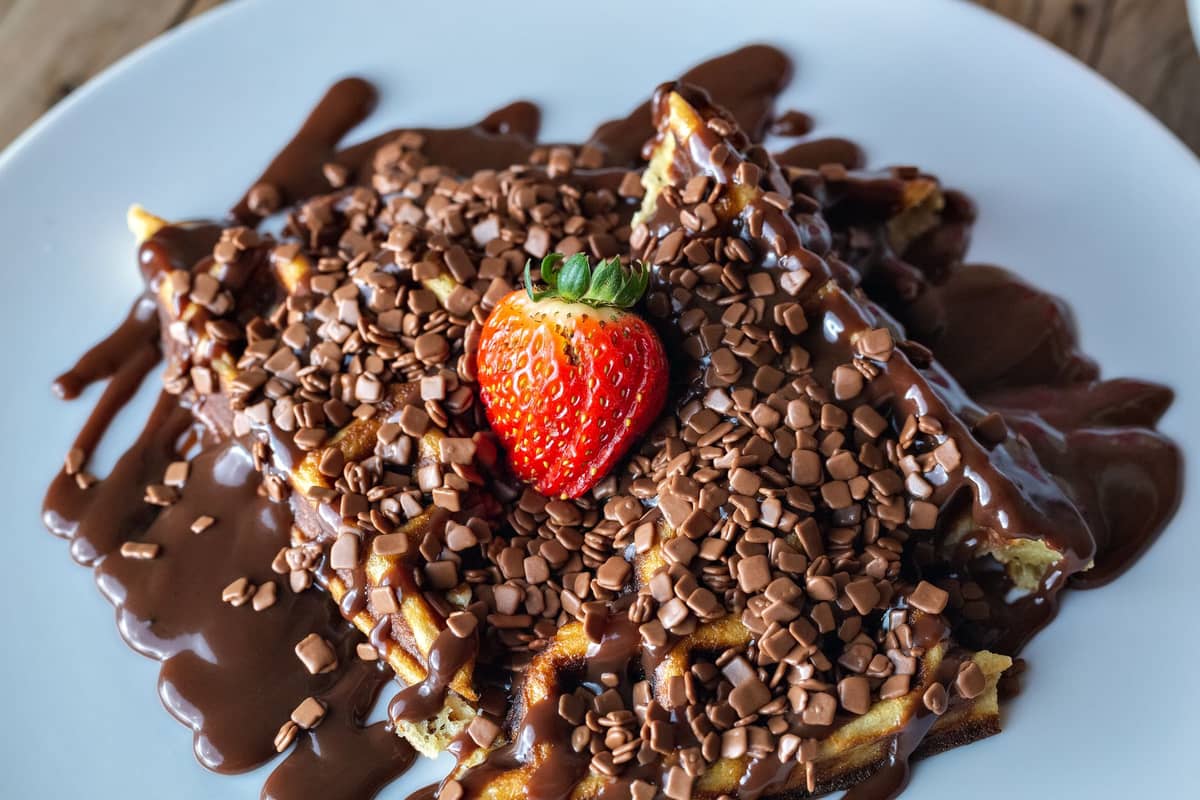 Waffle coberto com calda de chocolate e pedaços de chocolate, com um morango por cima, em um prato branco.
