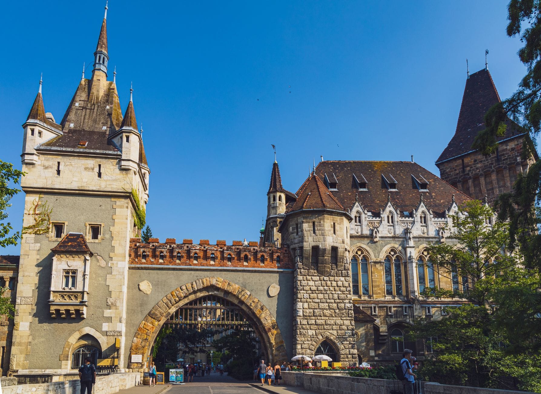 Blick auf den Eingang von Schloss Vajdahunyad im Budapester Stadtpark mit Menschen, die unter einem klaren blauen Himmel den Weg entlanggehen.