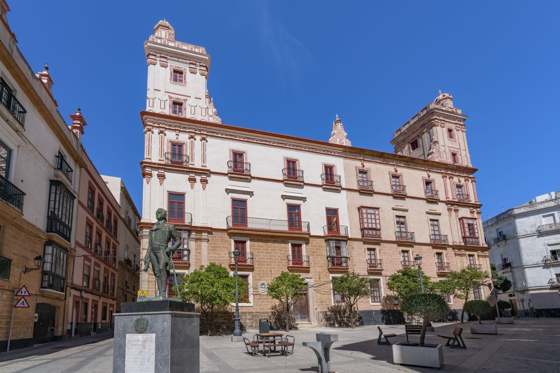 Grande edificio ornato con architettura storica in una piazza soleggiata, statua in primo piano e cielo azzurro sopra Cádiz, Spagna.