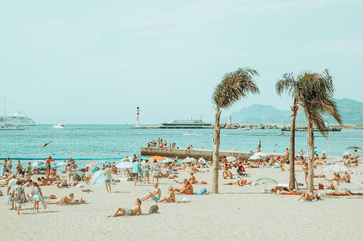 Concurrida playa de arena en Cannes con personas tomando el sol, palmeras, mar azul, yates y montañas al fondo