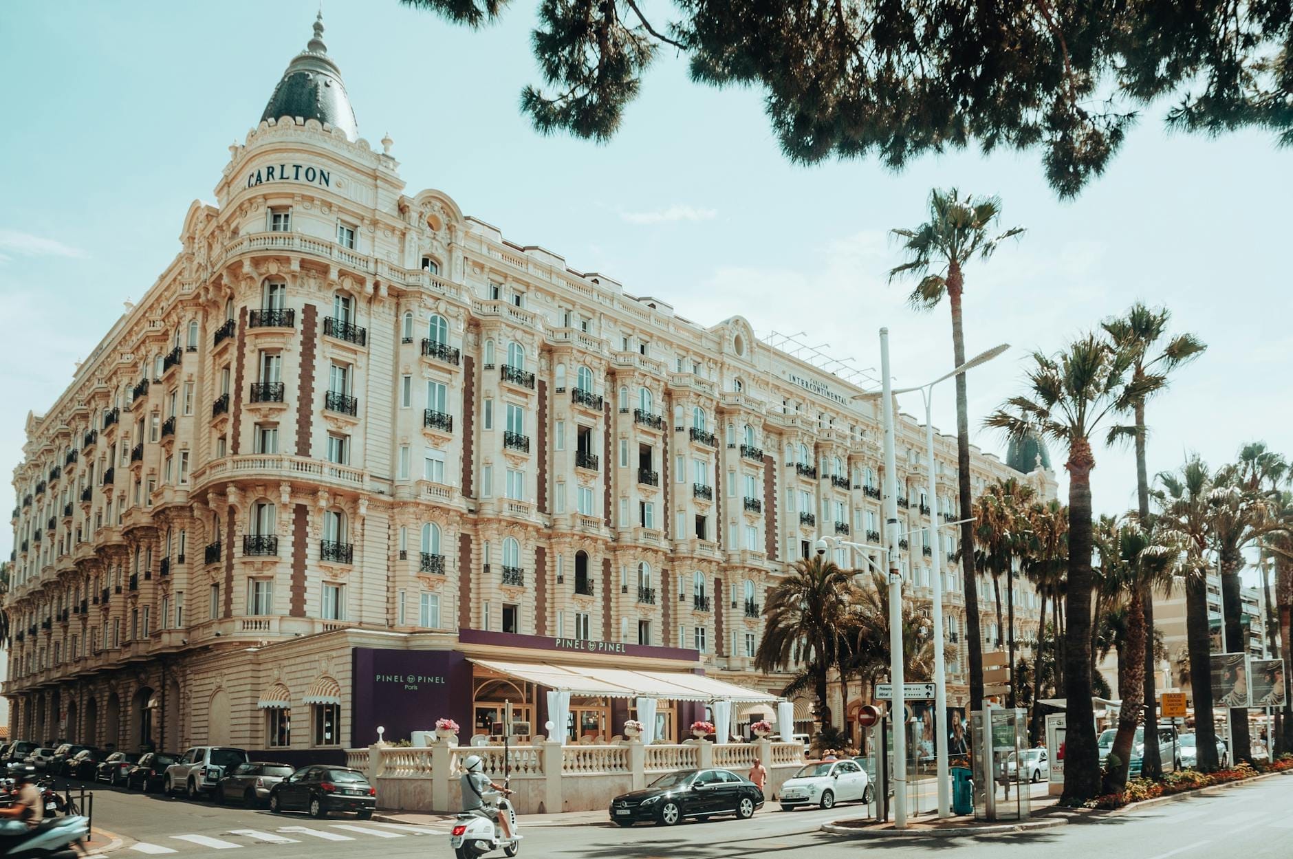 Vista elegante do histórico Hotel Carlton em Cannes, com palmeiras e pessoas na rua, representando a atmosfera icônica e glamourosa da cidade.