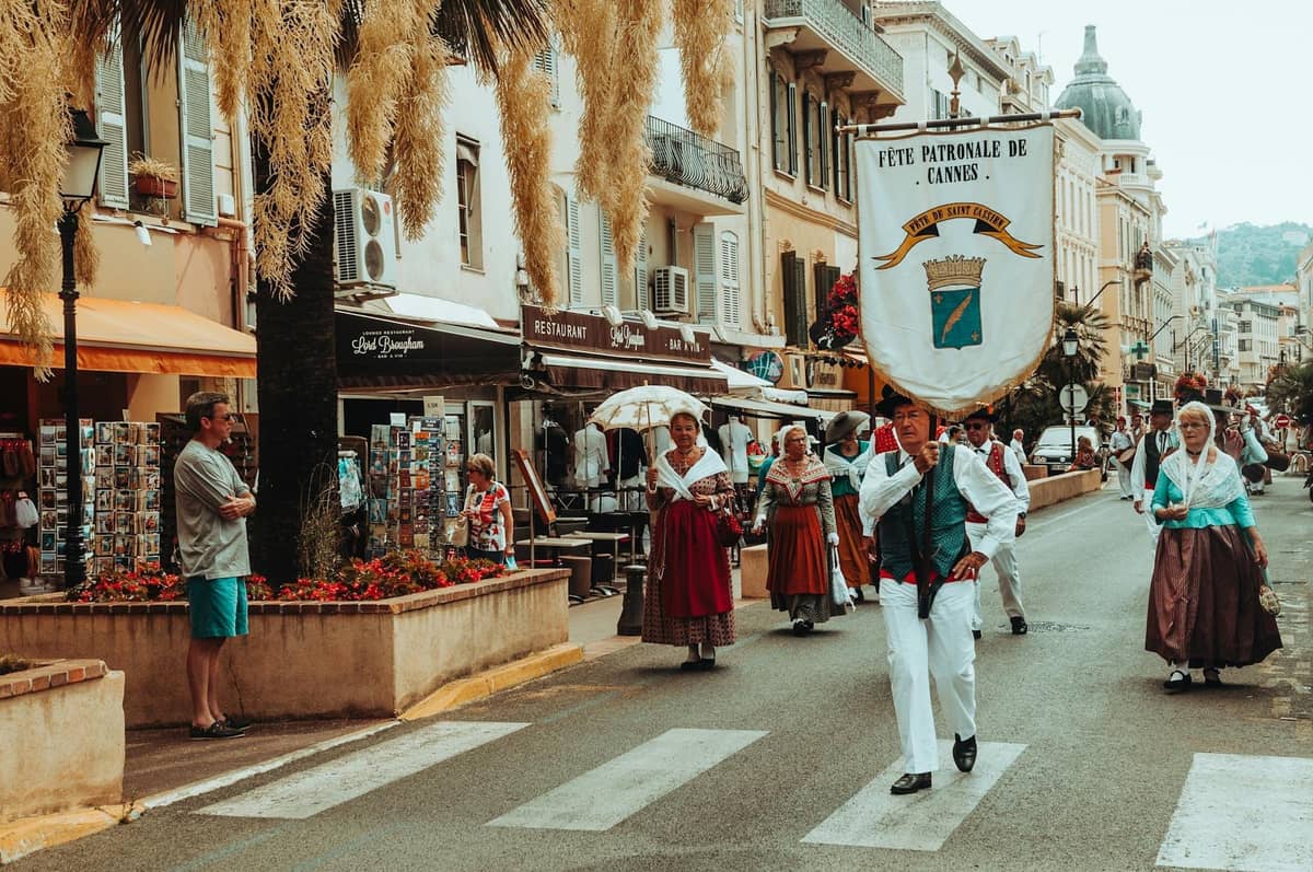 Personas con ropa tradicional desfilan por una animada calle de Cannes con cafés y tiendas a ambos lados.