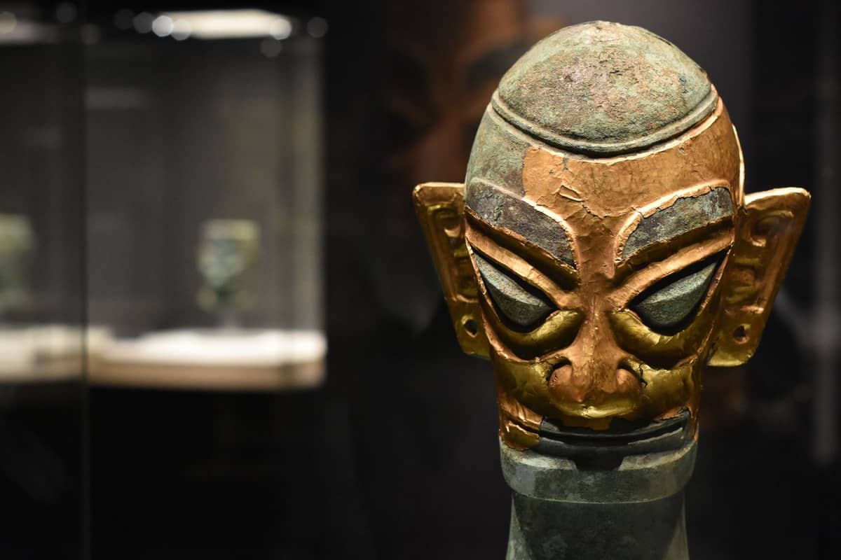 Nahaufnahme einer gold-bronzenen Sanxingdui-Maskenartefakt, die in einem Museum unter Scheinwerfern ausgestellt wird, mit anderen verschwommenen Artefakten im Hintergrund.