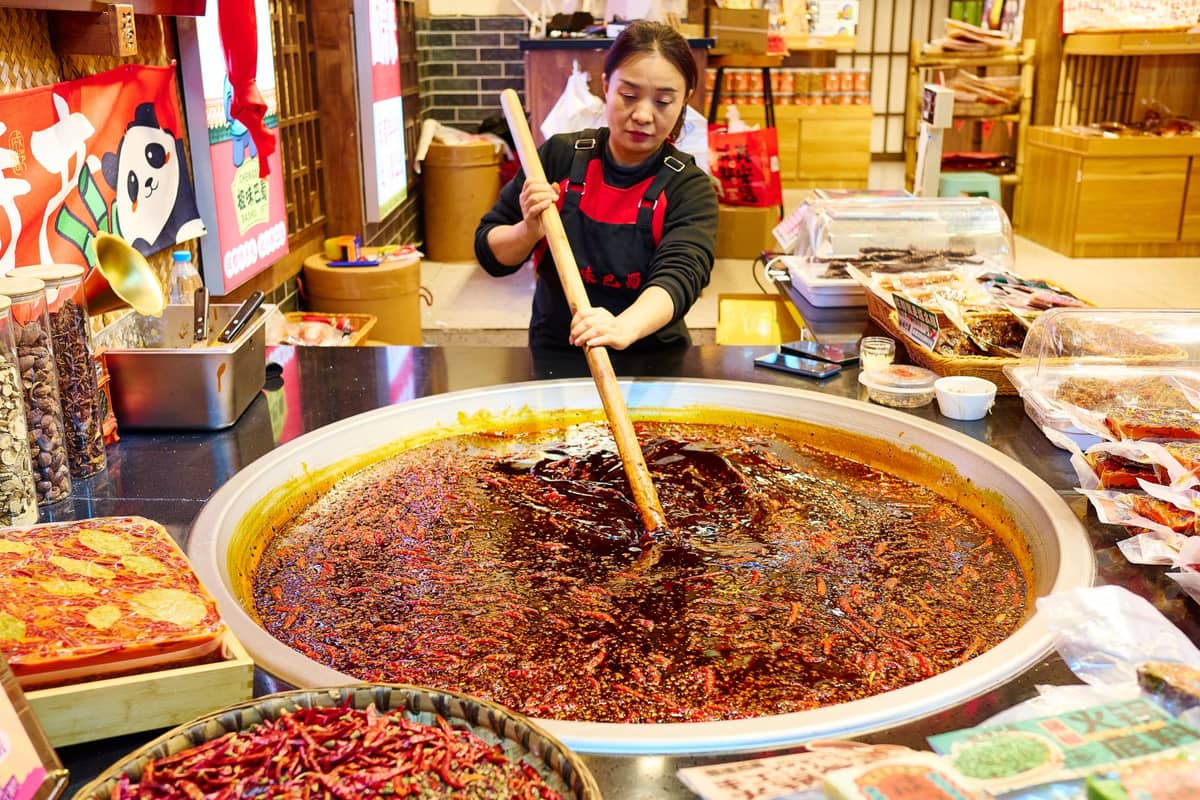 Eine Frau rührt in einem riesigen Topf mit Chiliöl und Gewürzen auf einem lebhaften Lebensmittelmarkt in Chengdu, umgeben von Sichuan-Snacks und roten Paprikaschoten.