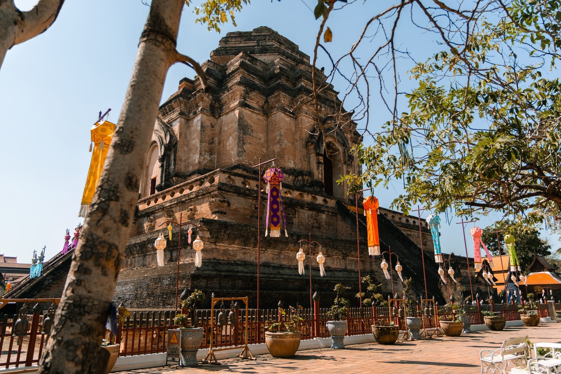 Antigua estupa de ladrillo del Wat Chedi Luang en Chiang Mai rodeada de coloridas linternas y banderas, árboles y cielo azul despejado durante el día.