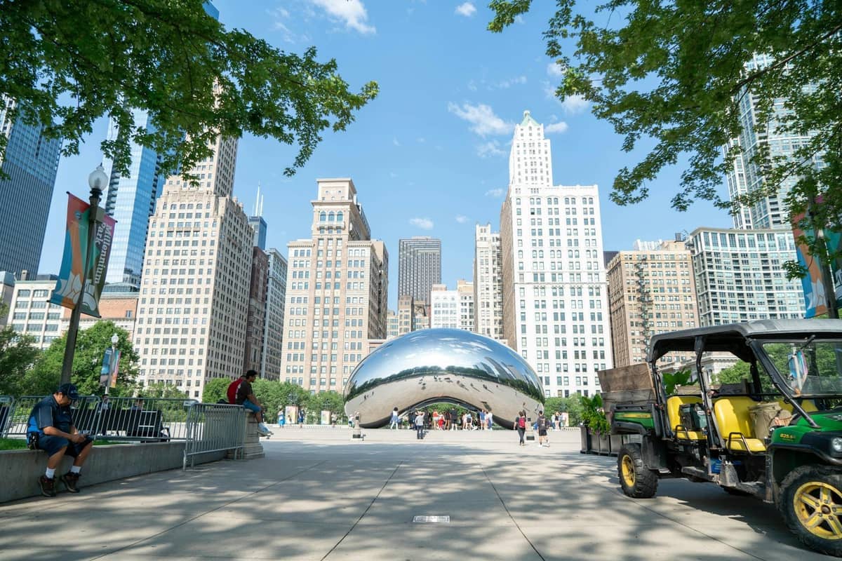 Cloud Gate Skulptur im Millennium Park mit umliegenden Wolkenkratzern und spazierenden Menschen an einem sonnigen Tag in der Innenstadt von Chicago.
