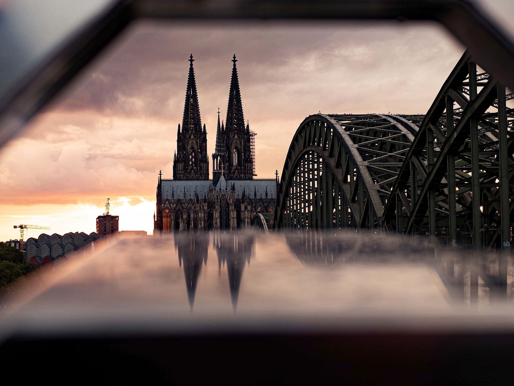 มหาวิหาร Cologne และสะพาน Hohenzollern ยามพระอาทิตย์ตก พร้อมเส้นขอบฟ้าของเมืองที่สะท้อนอยู่เบื้องหน้า ใต้เมฆยามเย็นที่งดงามตระการตา