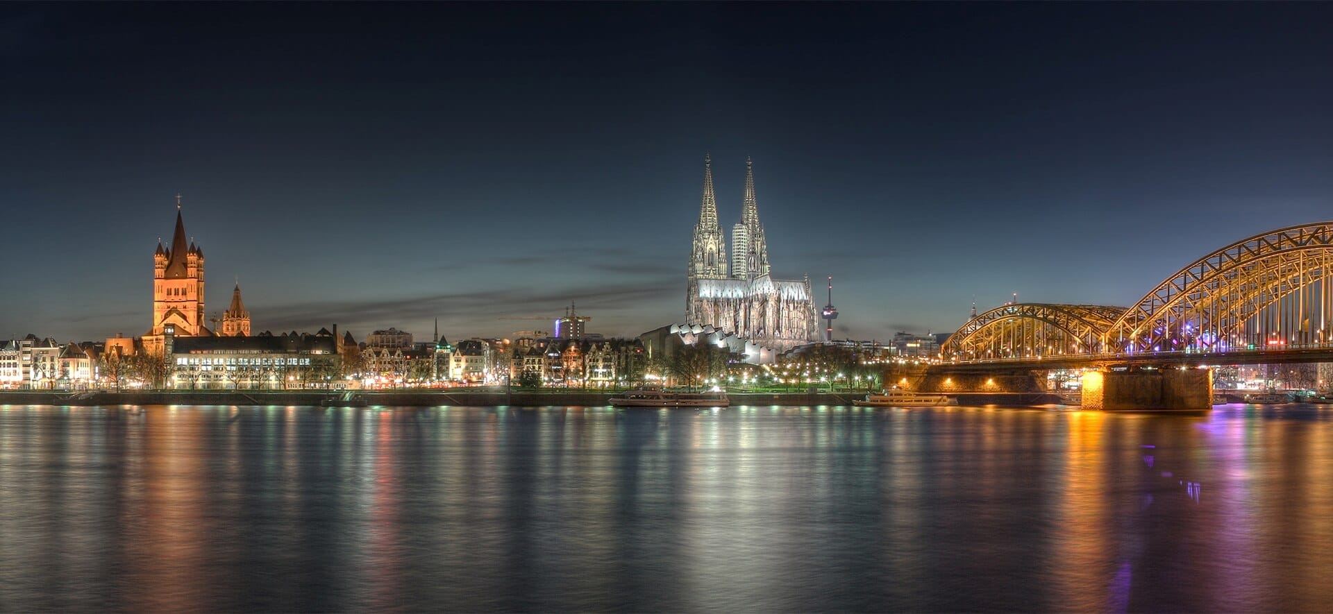 วิวพาโนรามายามค่ำคืนของเมืองโคโลญ นำเสนอมหาวิหารโคโลญที่สว่างไสว โบสถ์ Great St. Martin และสะพาน Hohenzollern ริมแม่น้ำไรน์
