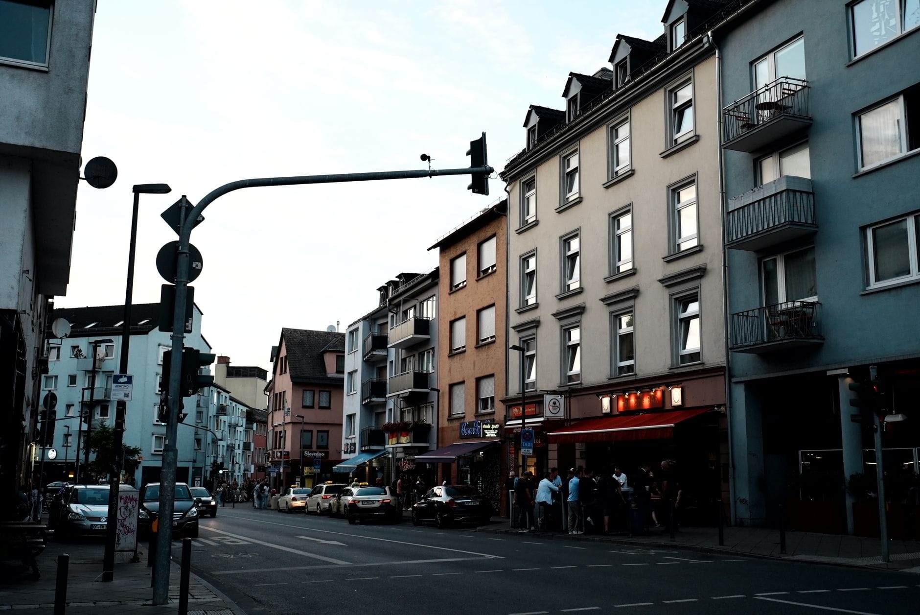วิวถนนในย่านเมืองที่คึกคักของ Cologne พร้อมผู้คน รถยนต์ ร้านกาแฟ และสถาปัตยกรรมผสมผสานที่เป็นเอกลักษณ์ของย่านเมืองที่ทันสมัย