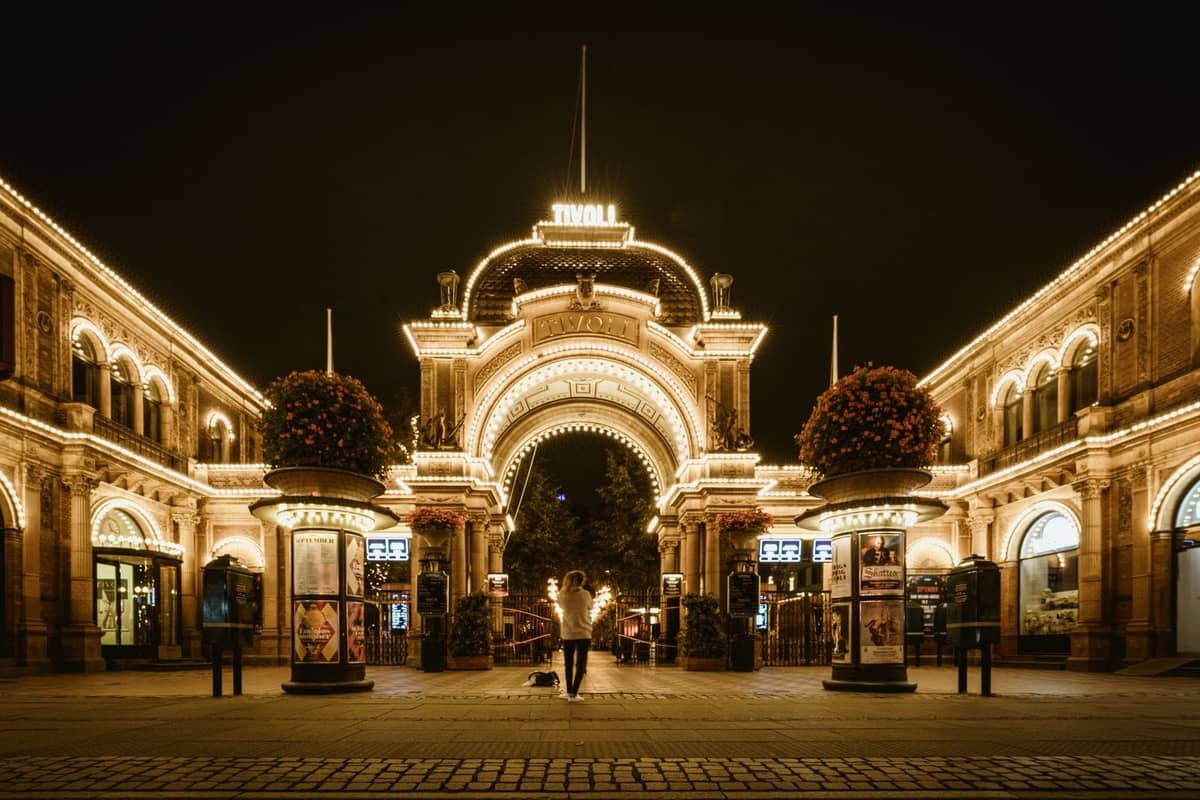 Entrada iluminada de Tivoli Gardens de noche con una persona de pie en el centro, rodeada de arcos ornamentados y luces decorativas.