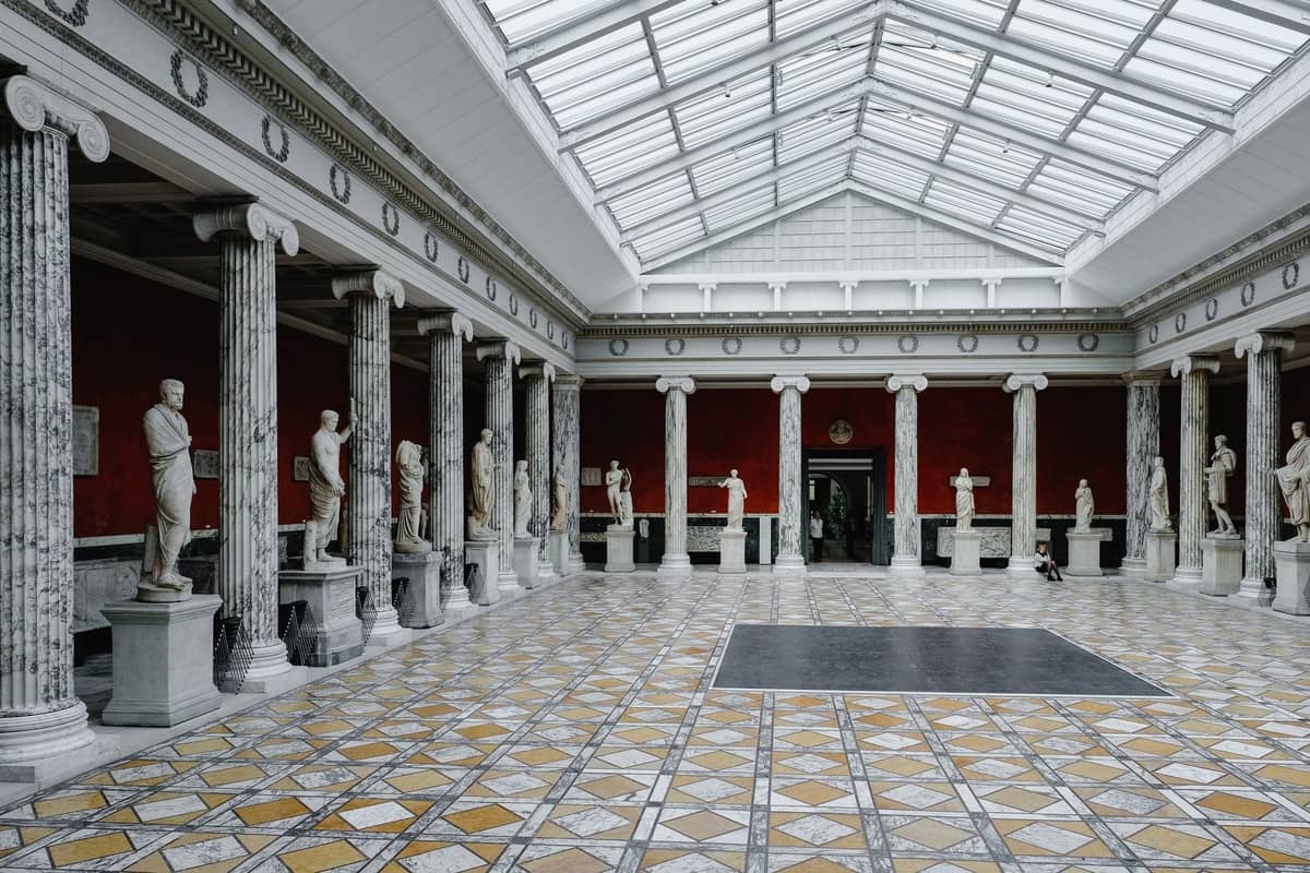 Gran sala de museo en Copenhague con columnas de mármol, paredes rojas, suelo con motivos decorativos y estatuas clásicas en exposición.