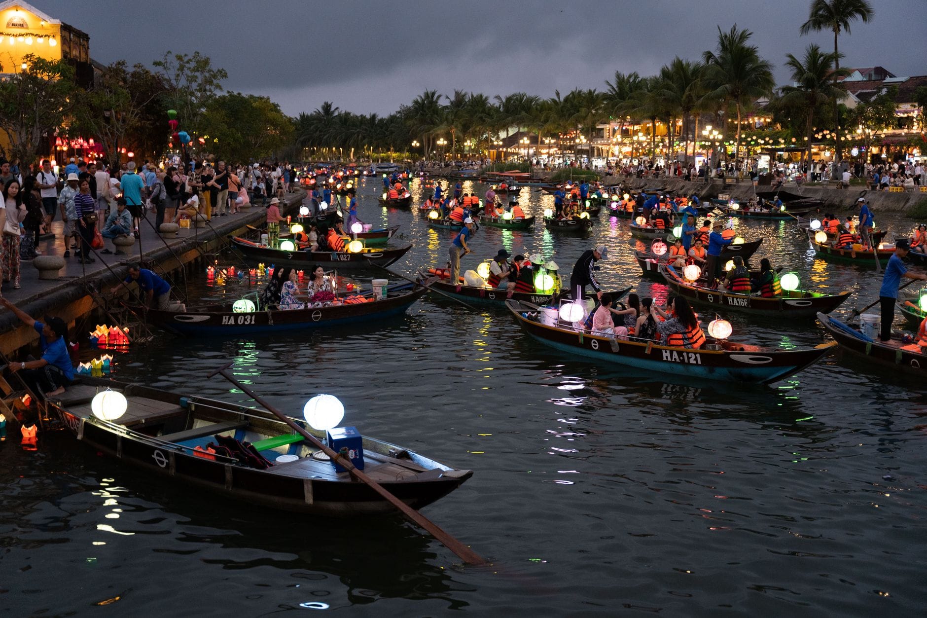 แม่น้ำที่คึกคักใน Hoi An ยามพลบค่ำ พร้อมเรือหลายลำที่บรรทุกโคมไฟและนักท่องเที่ยว มีต้นปาล์มและอาคารริมน้ำที่สว่างไสวเป็นฉากหลัง