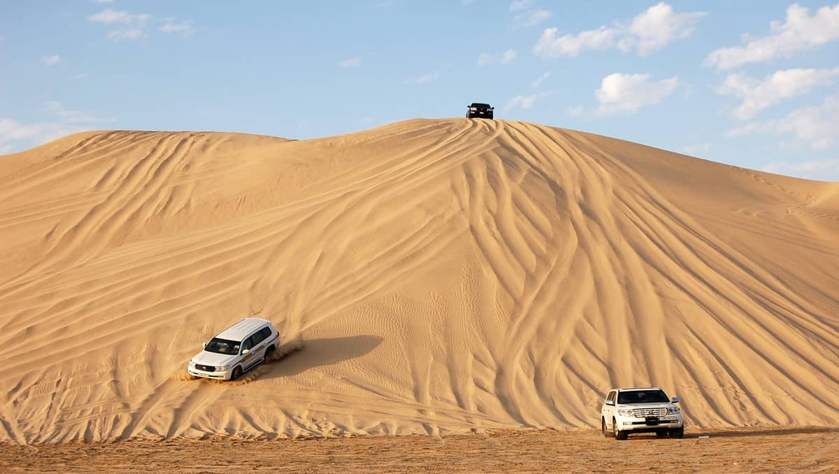 Drei Geländefahrzeuge fahren unter blauem Himmel über große goldene Sanddünen und zeigen das Abenteuer in der Wüstenlandschaft Katars.